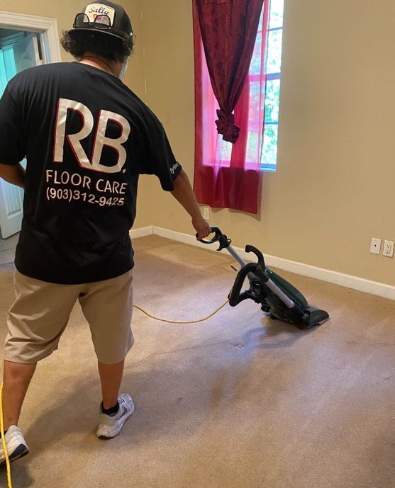 Worker vacuuming beige carpet in a room with white walls and a window with red curtains.