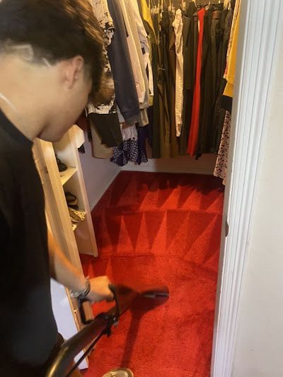 Person vacuuming a red carpeted closet. Clothes hang in the background.