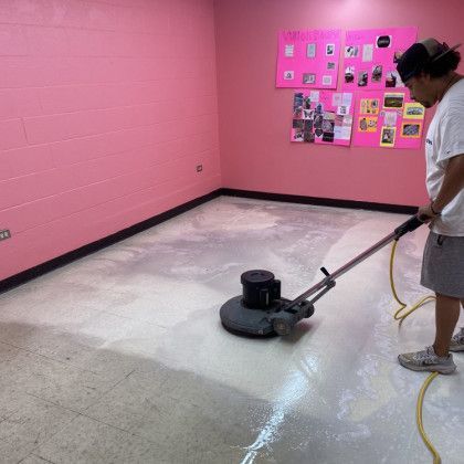 Person cleaning a light-colored floor with an industrial floor buffer in a pink-walled room.