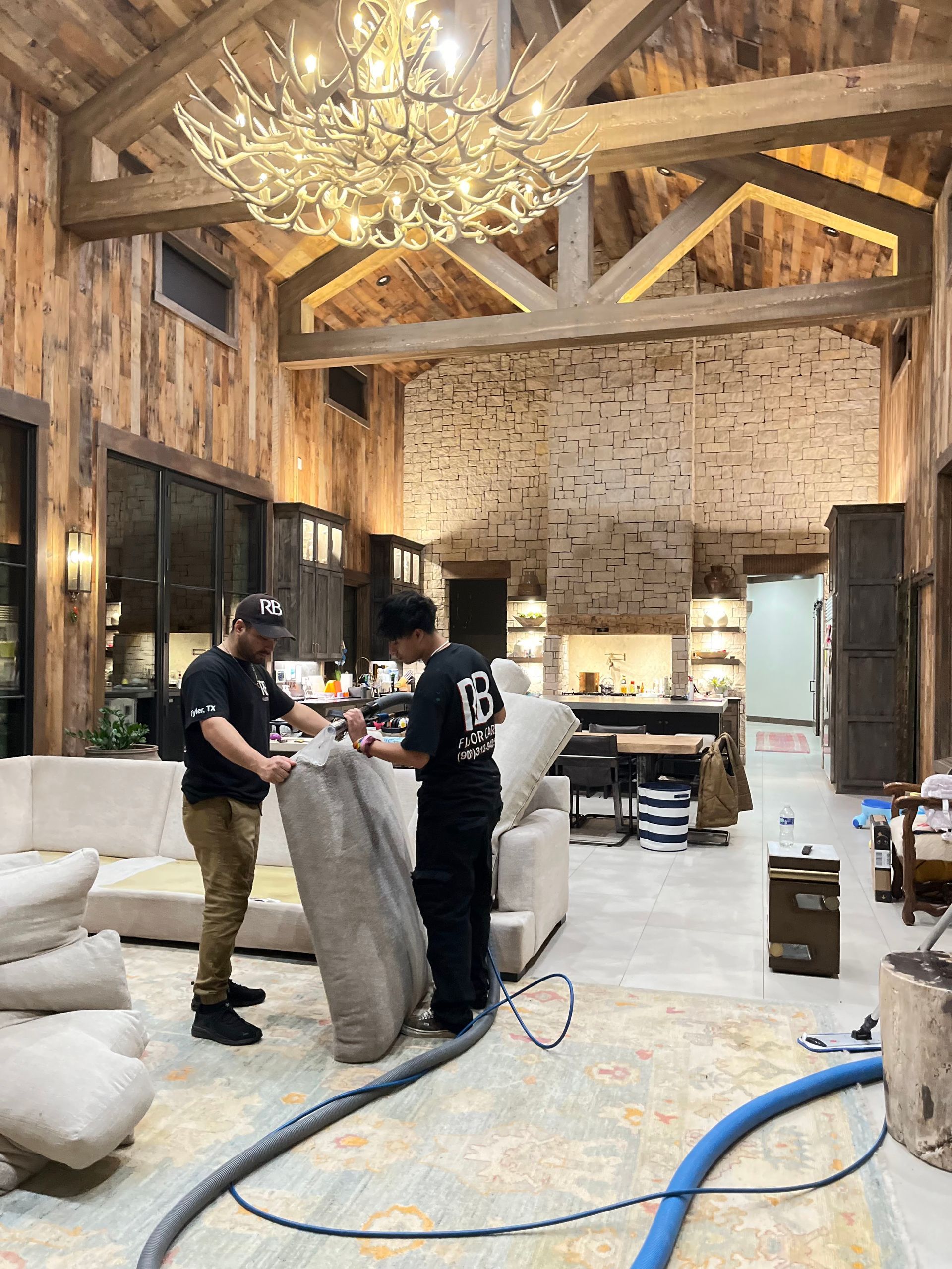Two workers moving a large bag inside a rustic barn-like room with exposed beams and a chandelier.