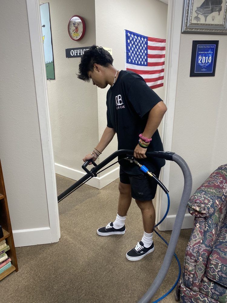 A young man vacuums a carpeted hallway; he wears a black shirt, shorts, and sneakers. An American flag hangs nearby.
