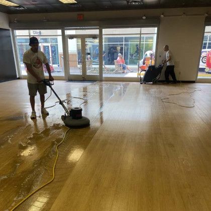 Worker polishing a wet wooden floor in a bright lobby with glass doors and a power cord