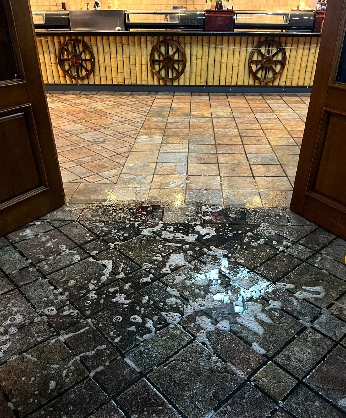 Puddled wet floor inside restaurant entrance, with open doors, decorative bamboo wall and wheel designs.