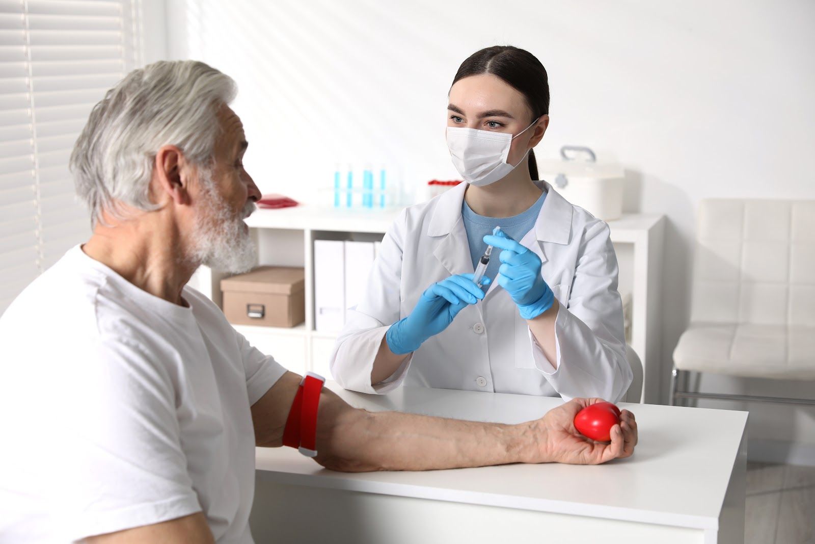 Doctor in mask and gloves examines an older patient’s arm in a clinic, with a red apple on the table.