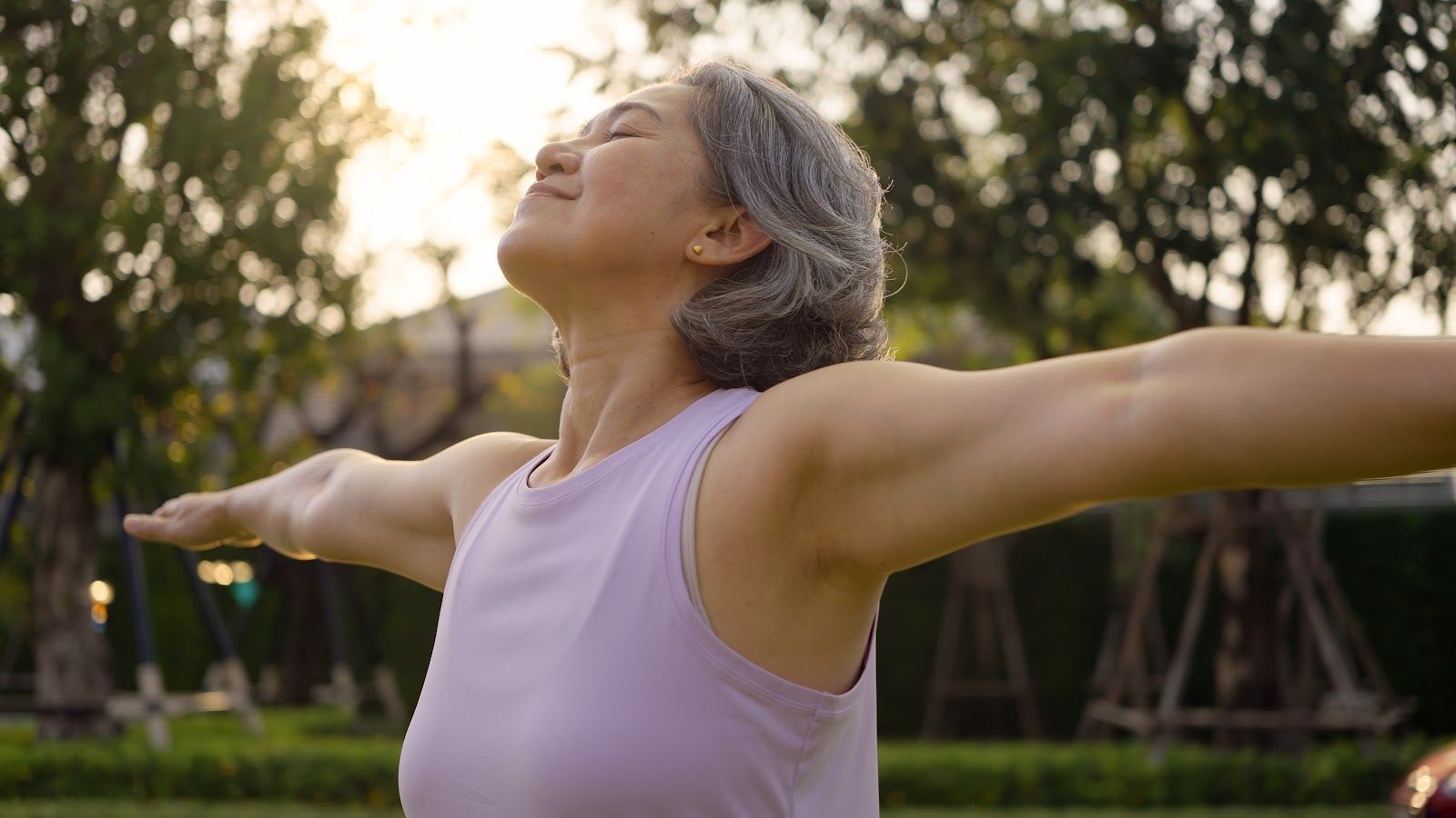 Woman in a lavender top stretching outdoors at sunset with arms outstretched and eyes closed