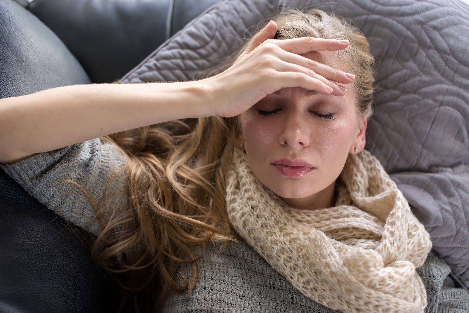 Woman reclining on a couch with hand on her forehead, looking unwell, wrapped in a knit scarf