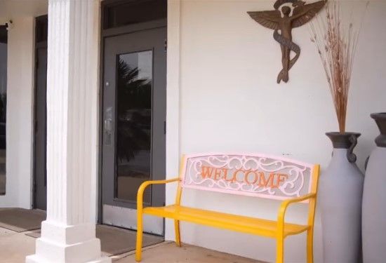 Yellow bench with a “Welcome” pillow on a porch beside a door and decorative vases