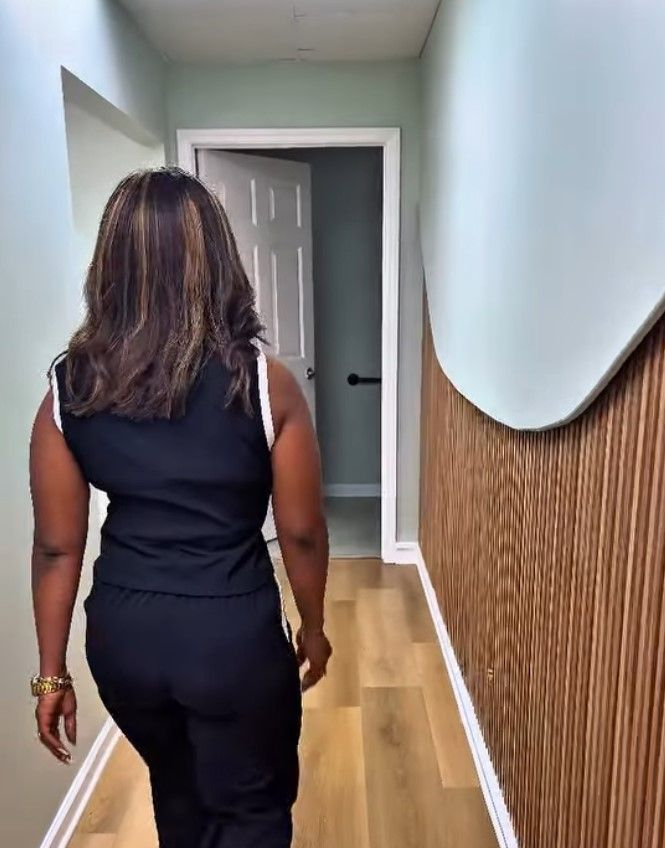 Person walking down a hallway with light wood flooring and white walls, seen from behind