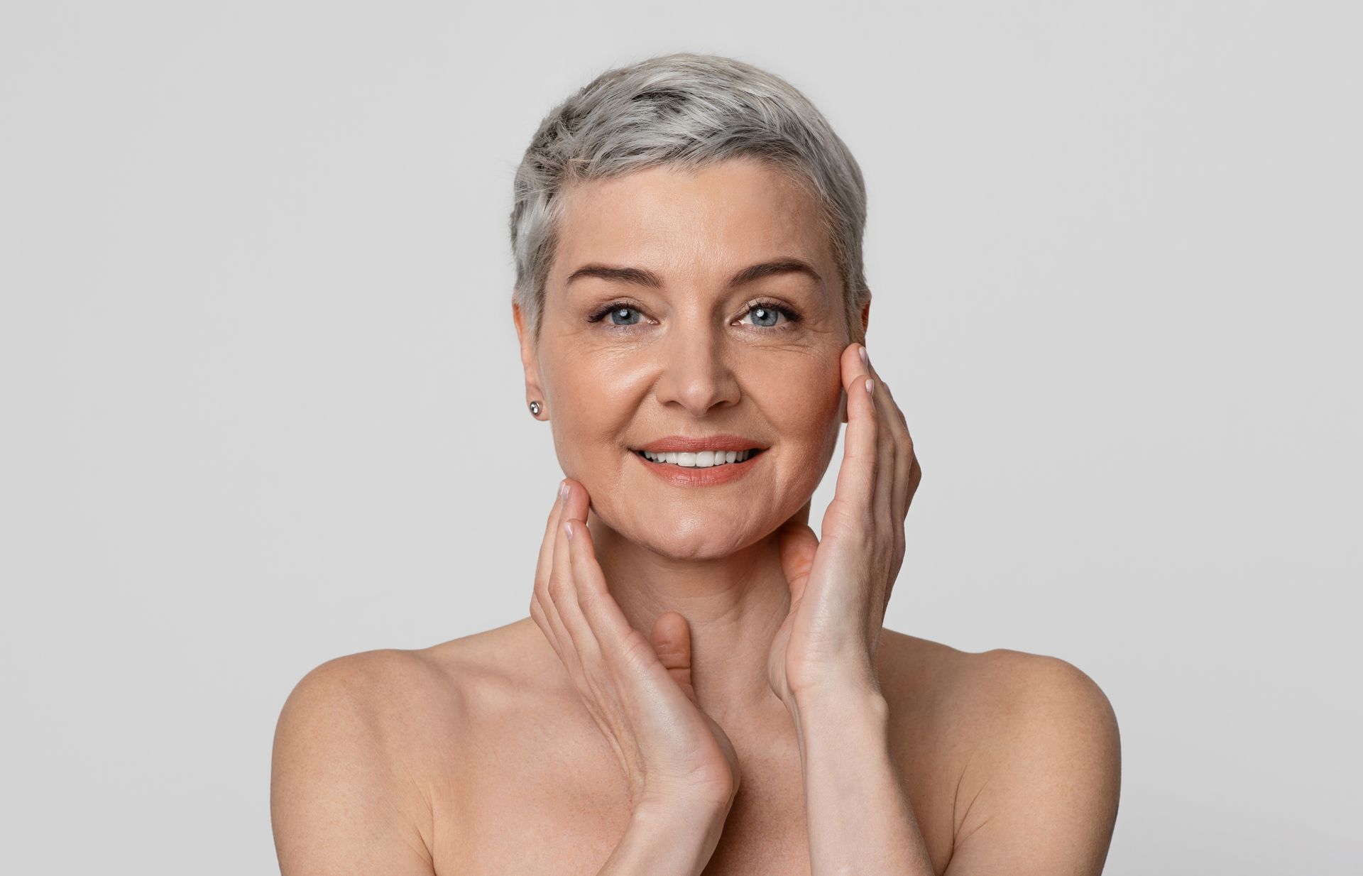 Smiling woman with short gray hair touching her face against a light gray background