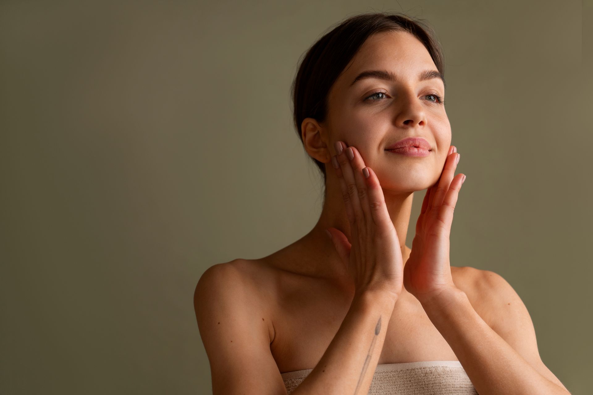 Woman gently applying skincare cream to her cheeks against a neutral background.