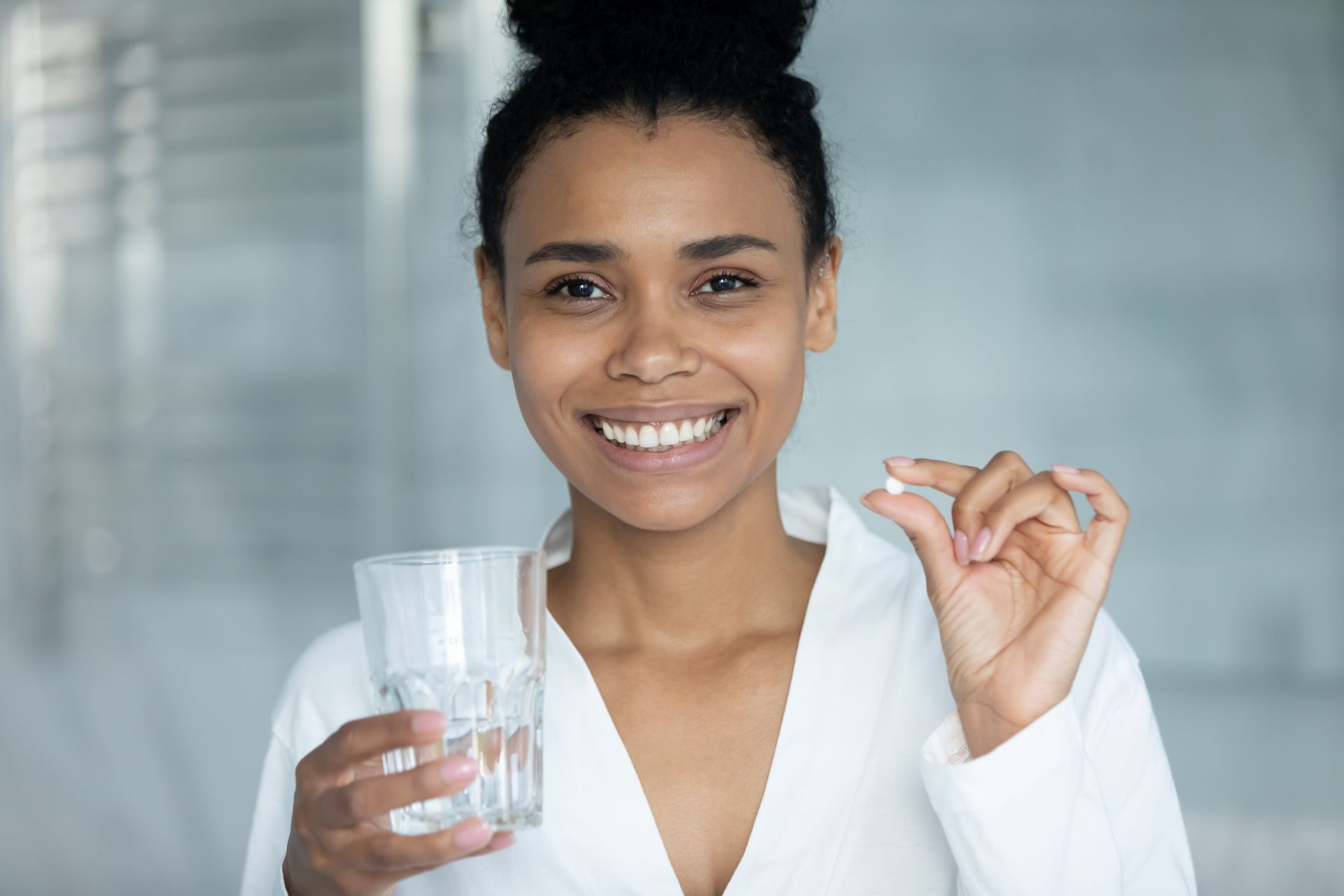 Smiling person in a white shirt holding a glass of water and a pill in a bright room