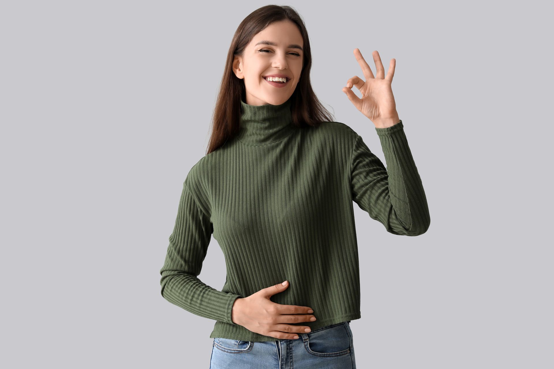 Smiling person in a green sweater making an OK gesture against a gray background
