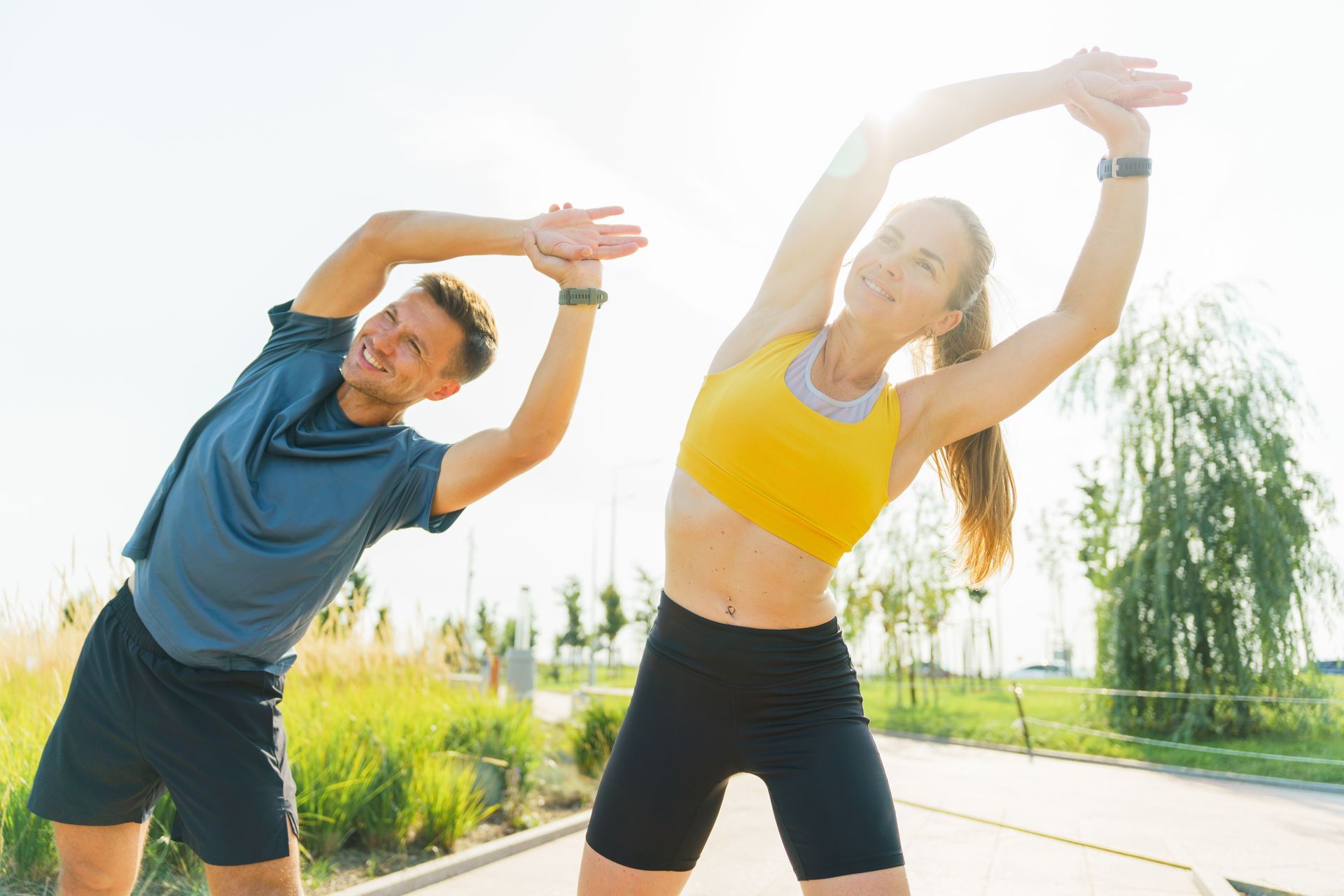 Two people stretching outdoors on a sunny path, wearing blue and yellow athletic clothes.