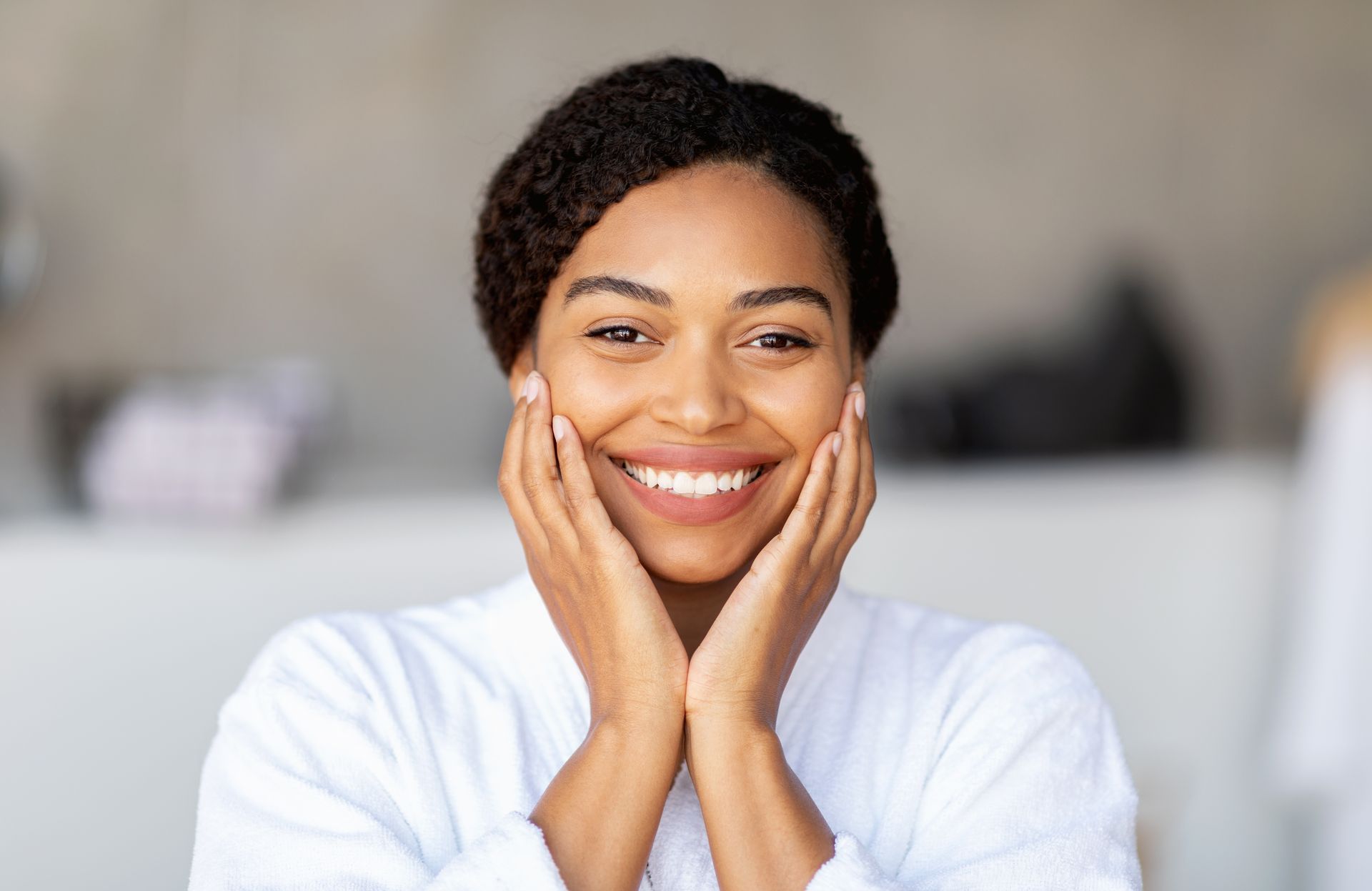 Smiling person in a white shirt resting hands on cheeks against a blurred indoor background