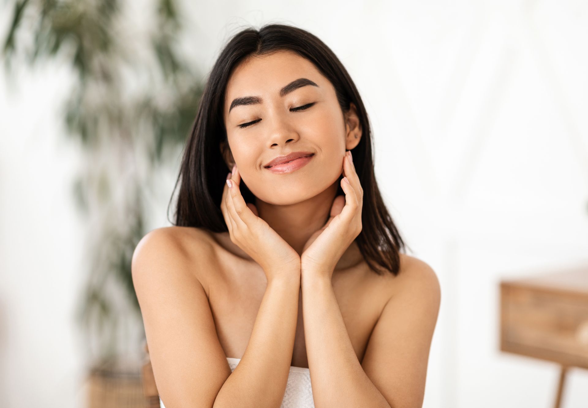 Smiling woman with closed eyes gently touching her face in a bright, softly lit room