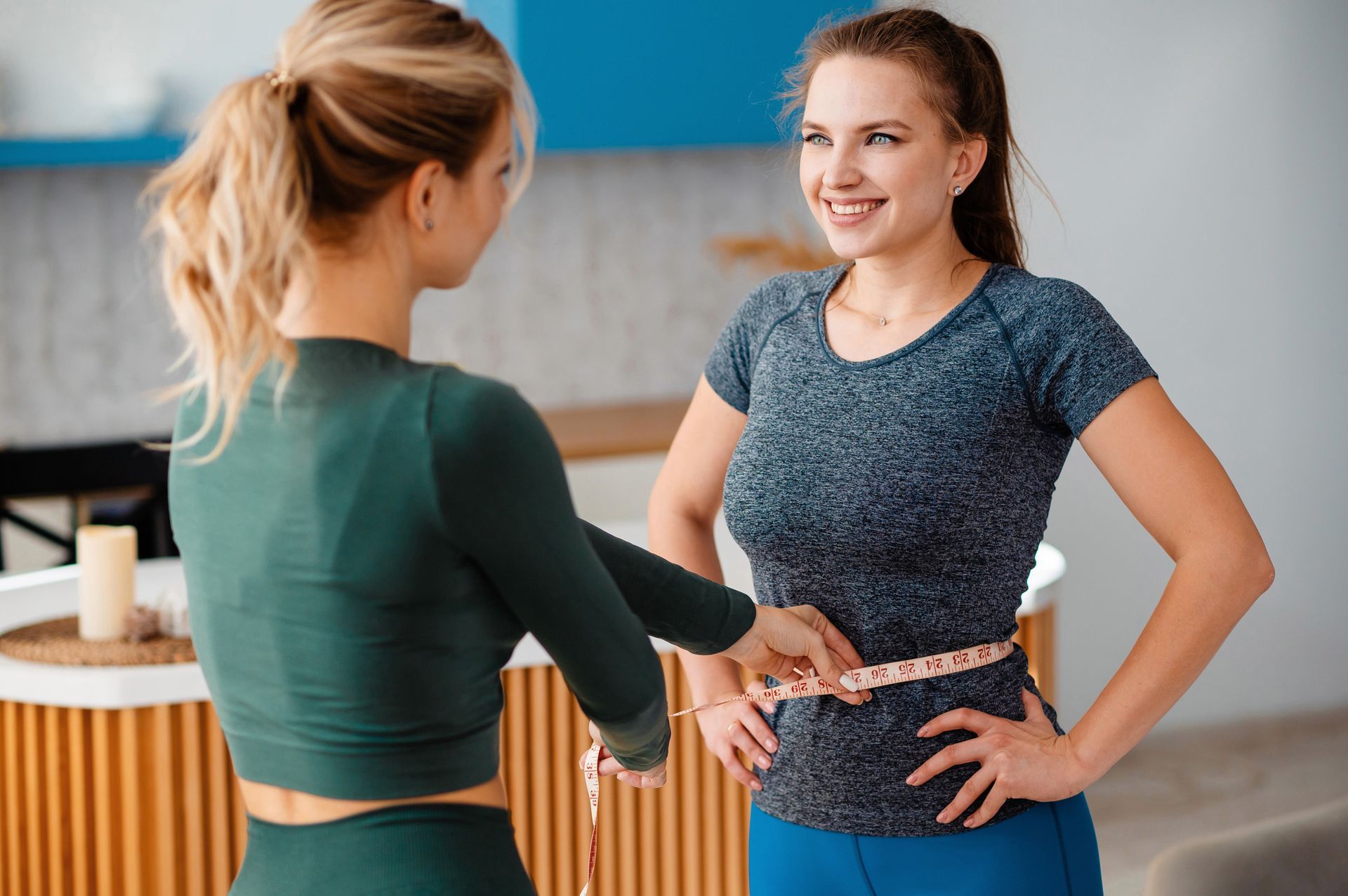 Two women in workout clothes talking in a gym, one smiling with hands on hips