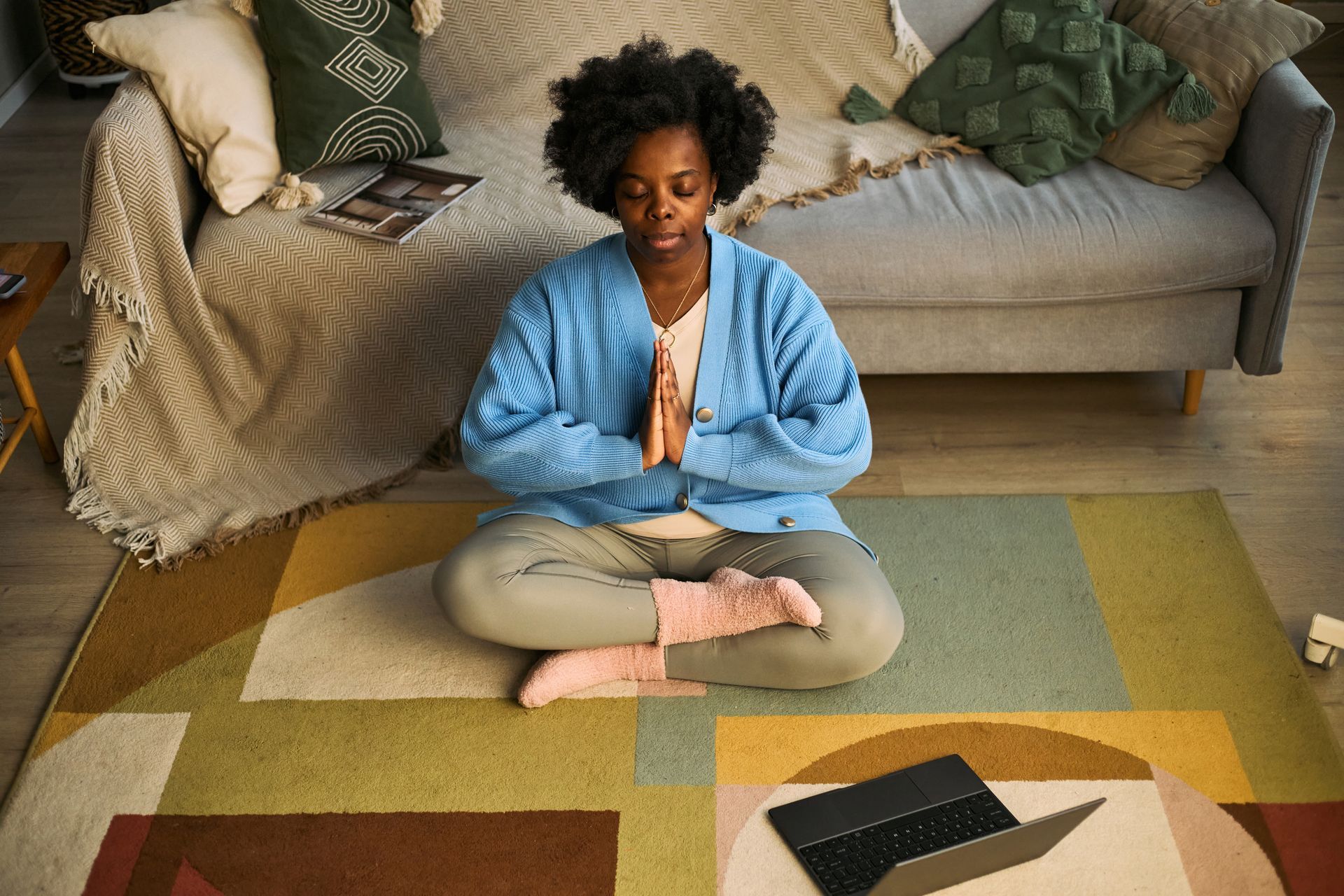 Person meditating cross-legged on a rug in a living room, with a couch and pillows behind.
