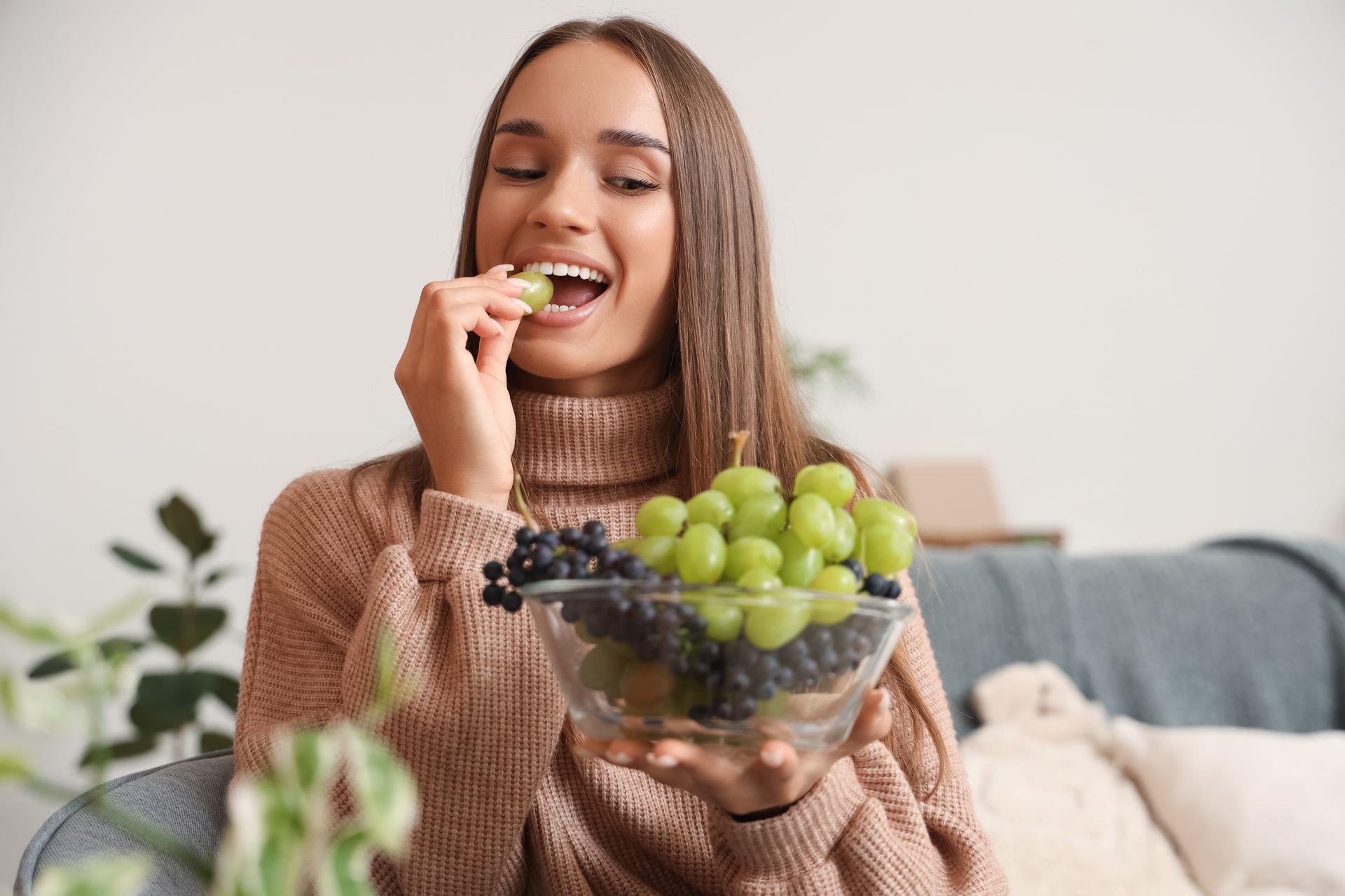 Woman eating a green grape from a bowl of fruit on a couch in a cozy living room
