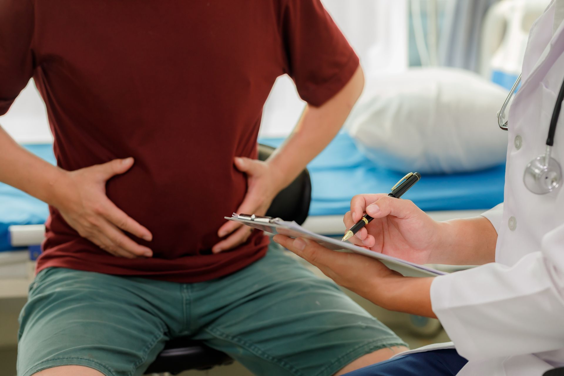 Doctor checking a seated patient’s abdomen in a clinic, with a pen and clipboard.