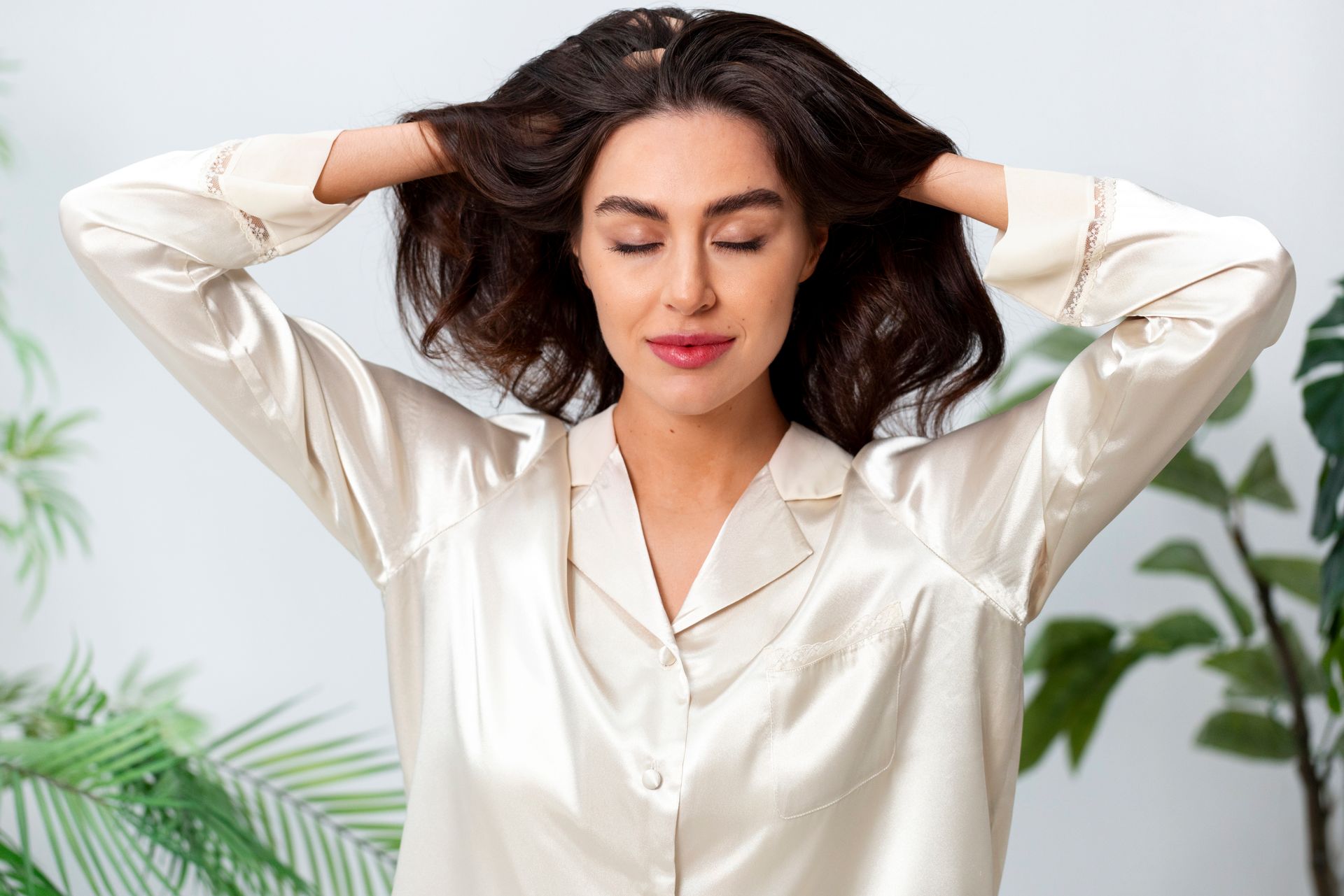 Woman in a white blouse with hands behind her head, eyes closed, in a bright indoor setting with plants.