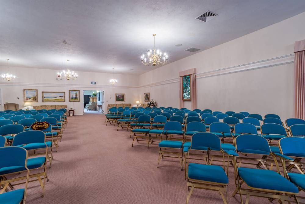 Empty auditorium with rows of blue chairs and a central aisle, lit by chandeliers and ceiling lights.