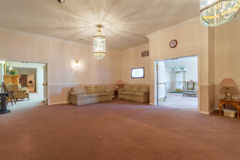 Spacious beige living room with brown carpet, sectional sofa, wall lights, and an open doorway to a kitchen.