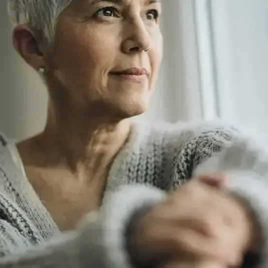 Thoughtful woman gazes out a window, wrapped in a gray knit sweater, with a blurred hand in the foreground