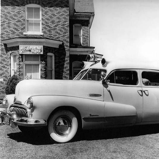 Vintage black-and-white car parked beside a brick house with a storefront sign.