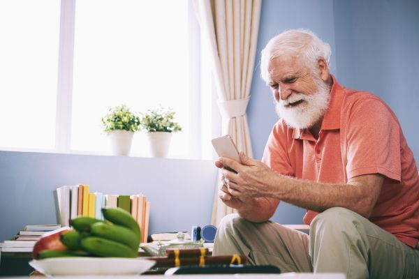 Older man in a coral shirt smiling at a smartphone in a bright living room with fruit and books nearby