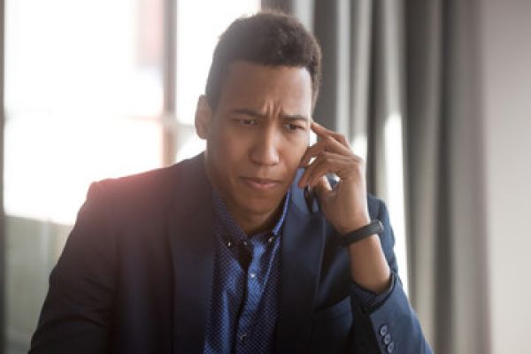 Man in a dark blazer speaks on a phone by a bright window, looking concerned.