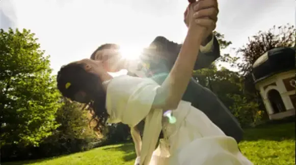 Couple dancing and leaning back in a sunlit garden, with trees and a house in the background