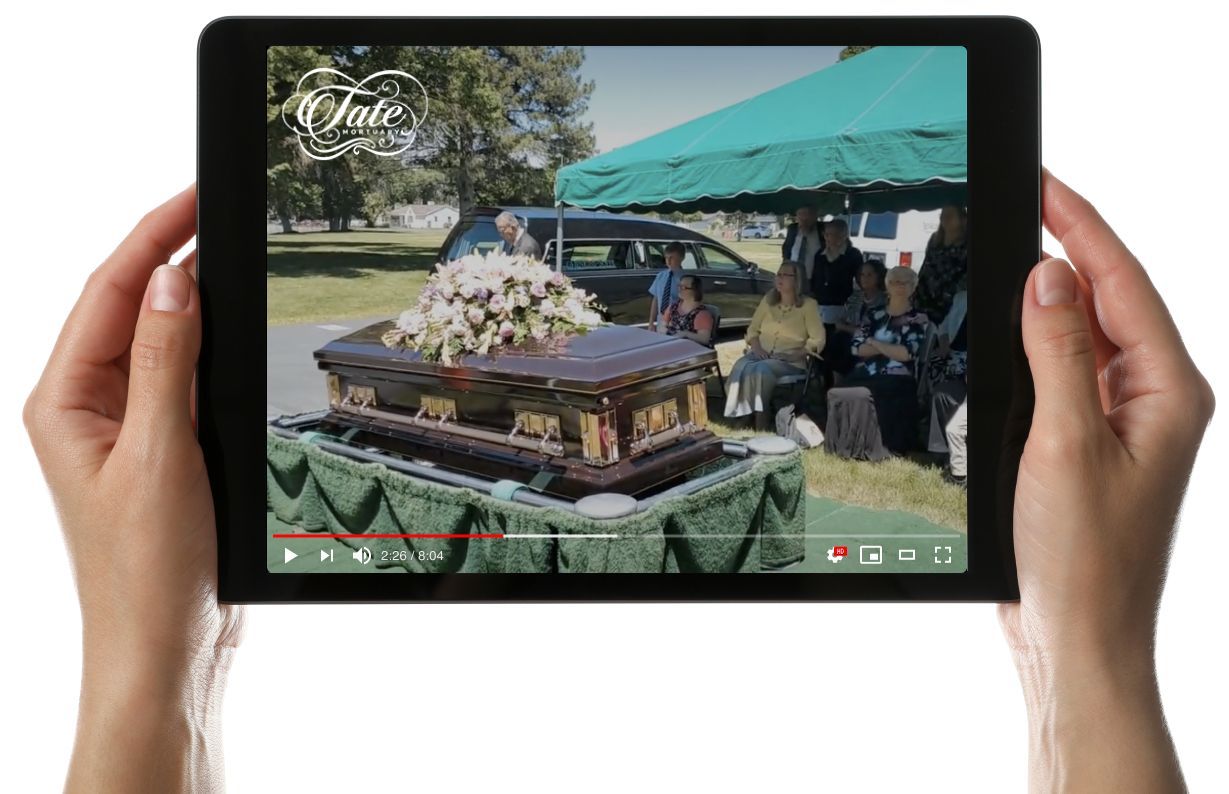 Hands holding a tablet playing a video of a flower-covered coffin at an outdoor funeral.