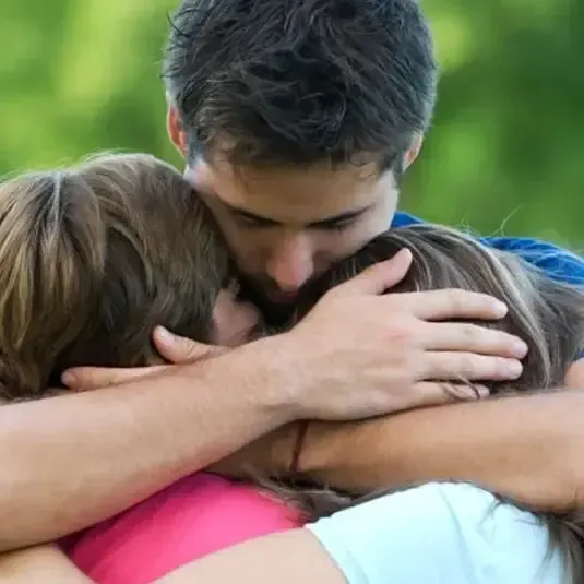 Three people hugging closely outdoors on grass, with heads together in a group embrace.