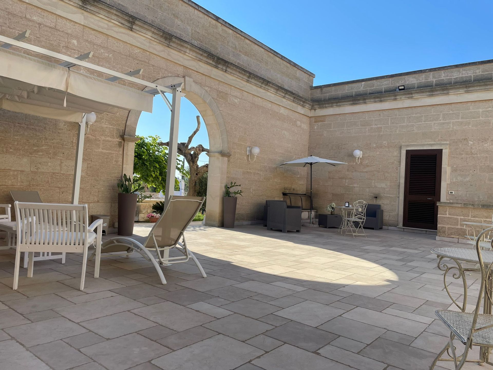 Patio esterno con muri in pietra, mobili e un ombrellone nelle giornate di sole.