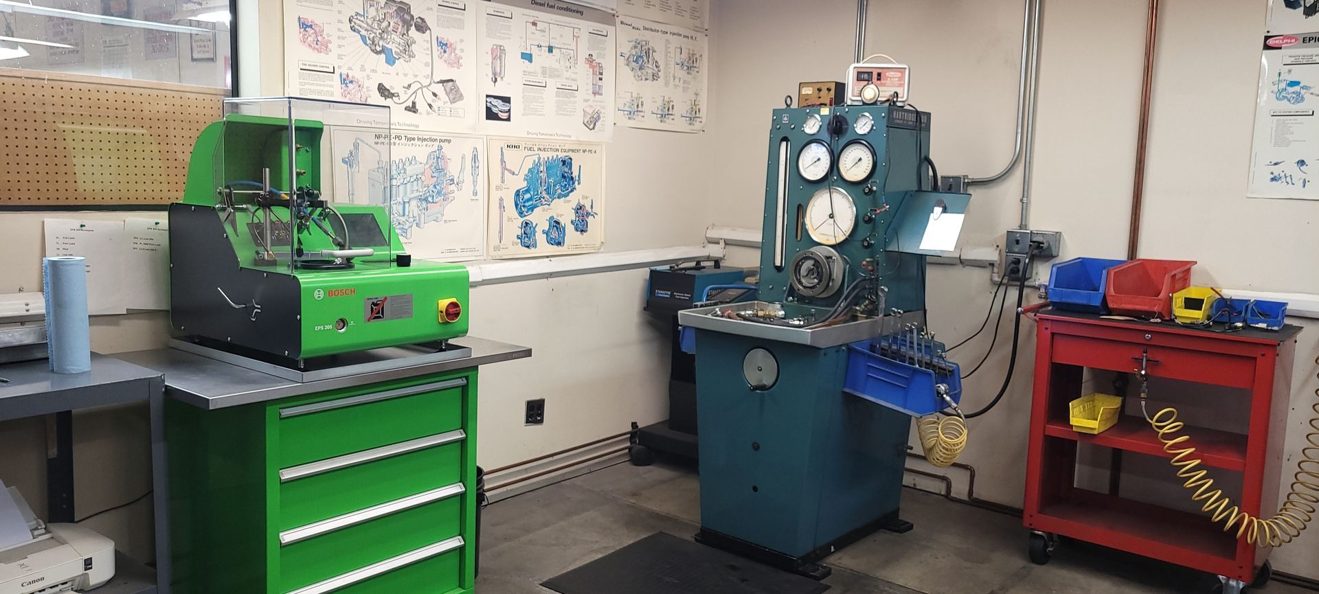 Industrial workshop with green tool chest, blue machine, and red cart against a wall of drawings
