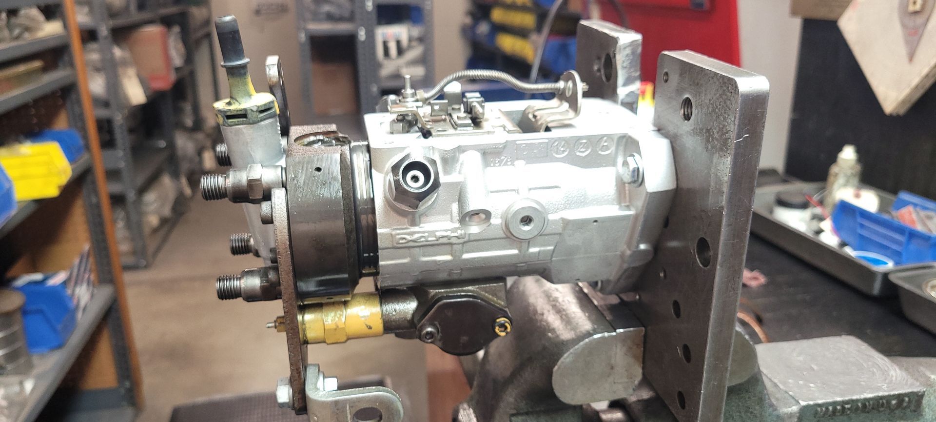 Silver mechanical part clamped in a workshop vise, surrounded by tools and equipment.