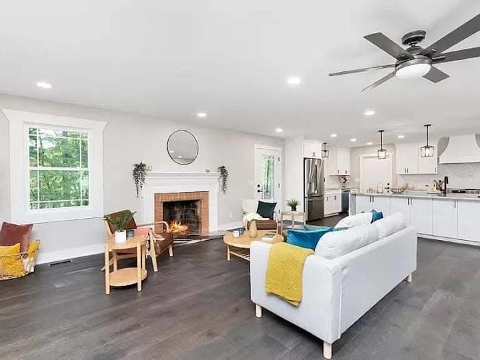 Open-concept living room with fireplace, white walls, dark wood floors, and kitchen in the background.