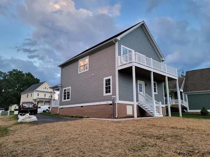 Two-story house with gray siding, white trim, and a balcony. Brick base, driveway, and dry yard. Cloudy sky.