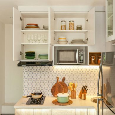 Bright white kitchen with open cabinets, microwave, stove, and wooden utensils on a countertop with patterned tile backsplash.