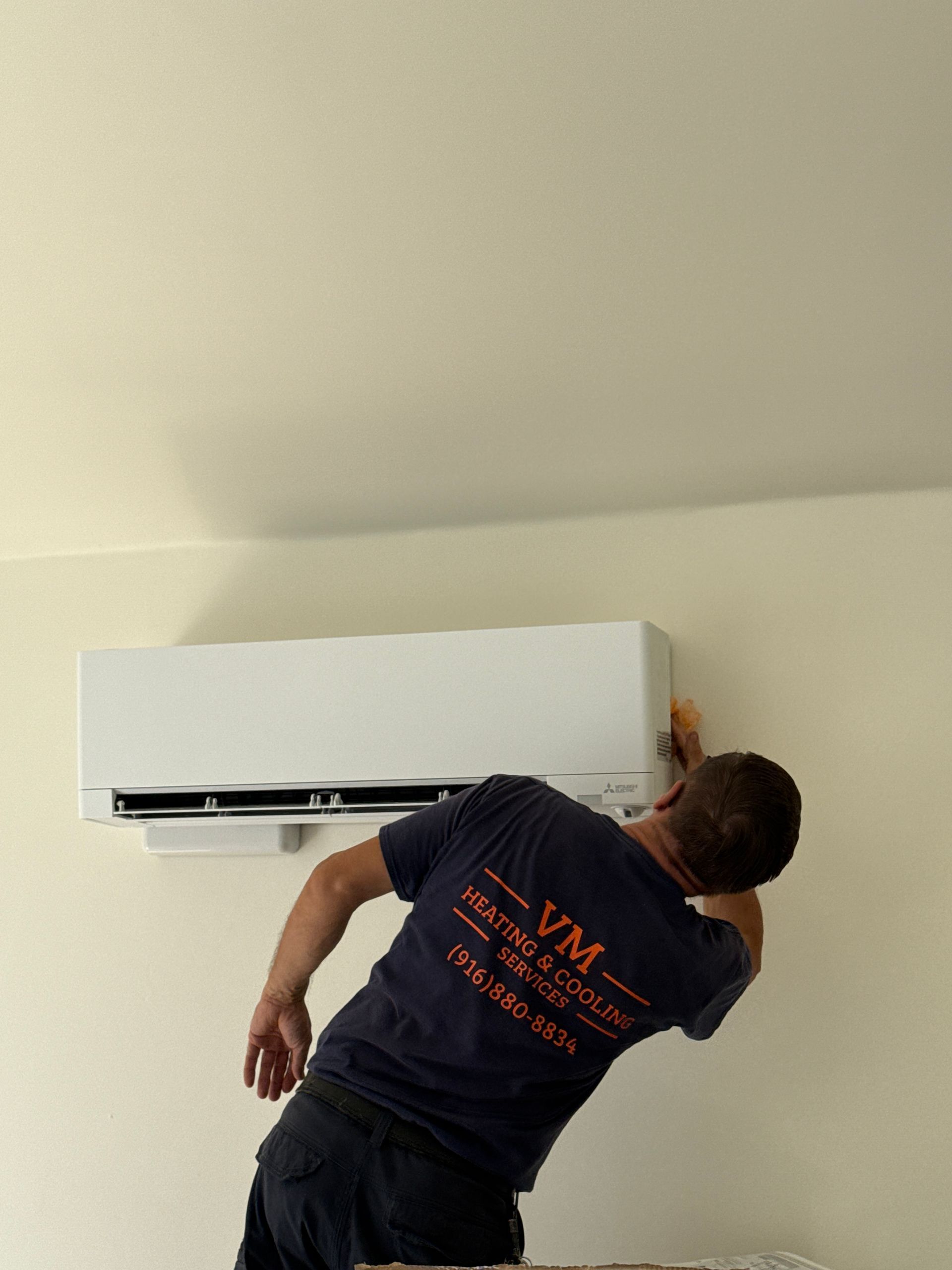 Person installing a wall-mounted air conditioner on a white wall, leaning up with tools.