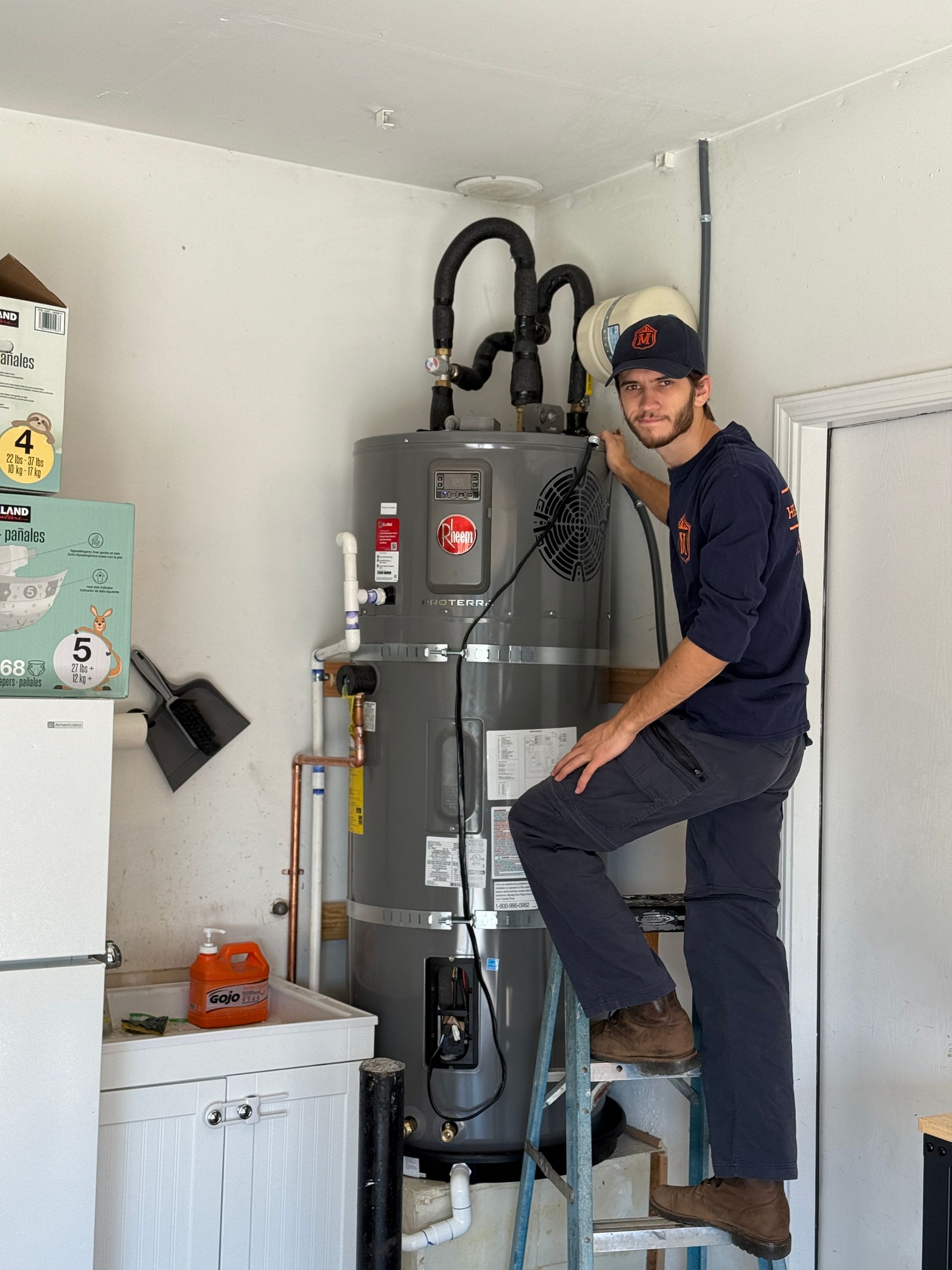 Worker standing on a ladder beside a large gray water heater in a garage laundry area