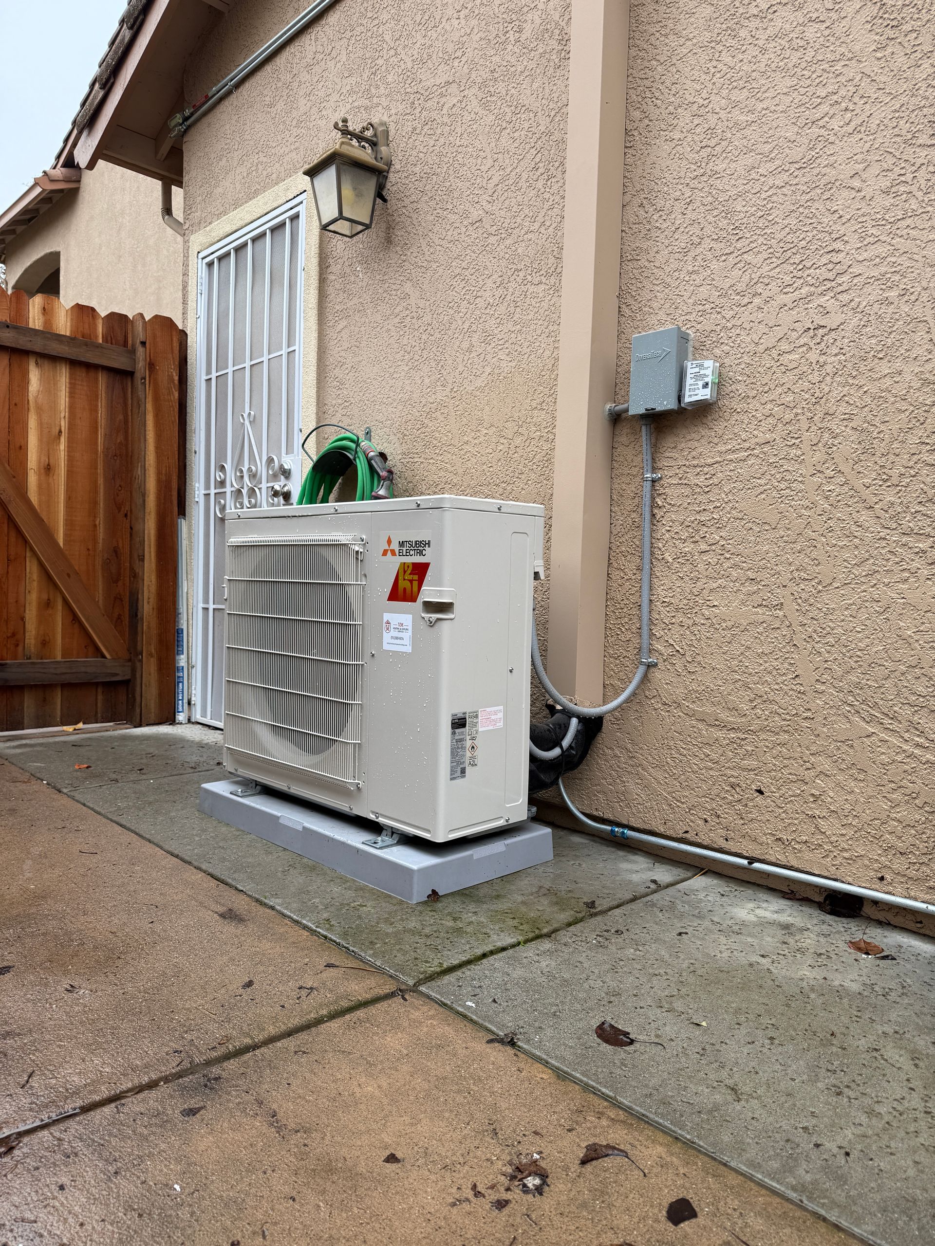 Outdoor HVAC unit beside a stucco house wall, next to a wooden gate and concrete walkway.