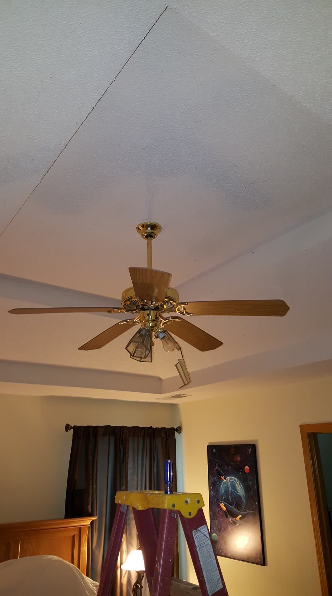 Ceiling with a crack, a ceiling fan, a ladder, and a framed art piece in a bedroom.