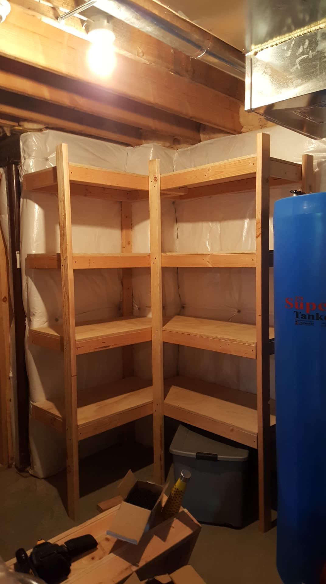 Wooden corner shelving in a basement; shelves filled with storage against concrete wall and water tank.