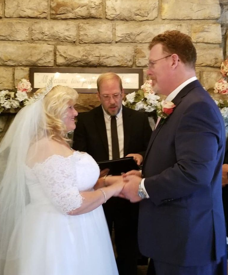 Bride and groom hold hands during their wedding ceremony, facing officiant in front of a stone wall.