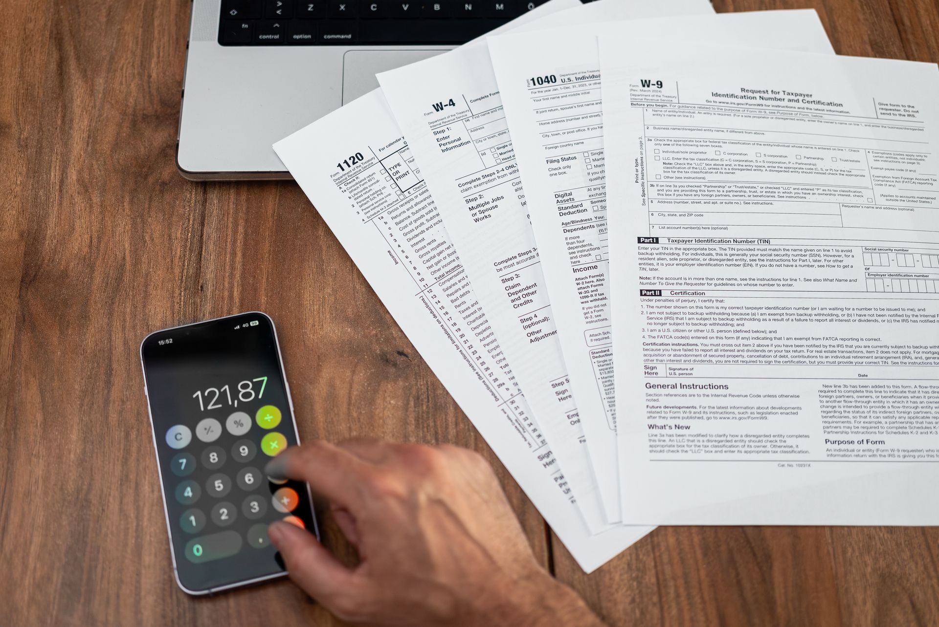 Hand using calculator on smartphone, tax forms, and laptop on wooden desk.