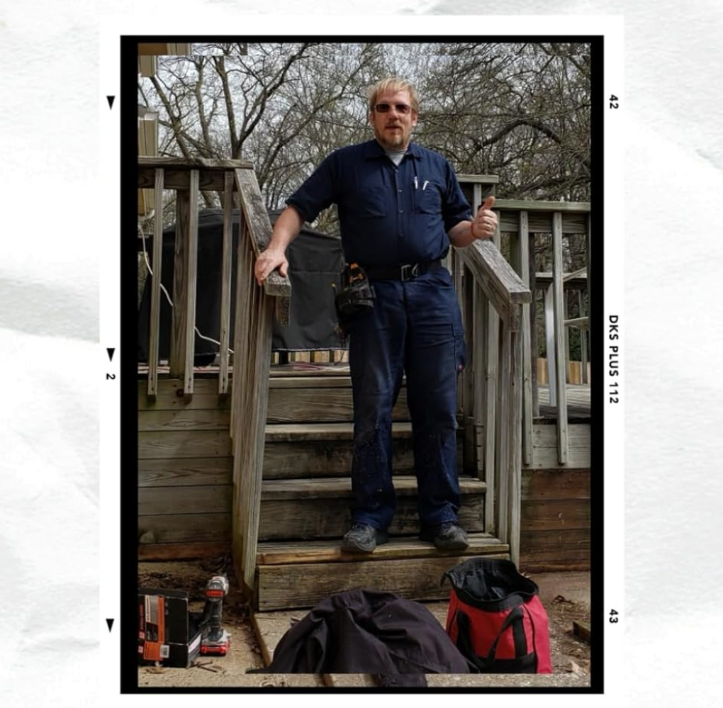 Man in work clothes on wooden stairs, thumbs up. Outdoor setting, tools nearby.