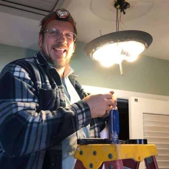 Man on a ladder, installing a light fixture; smiling, indoor, wearing work clothes and a headlamp.