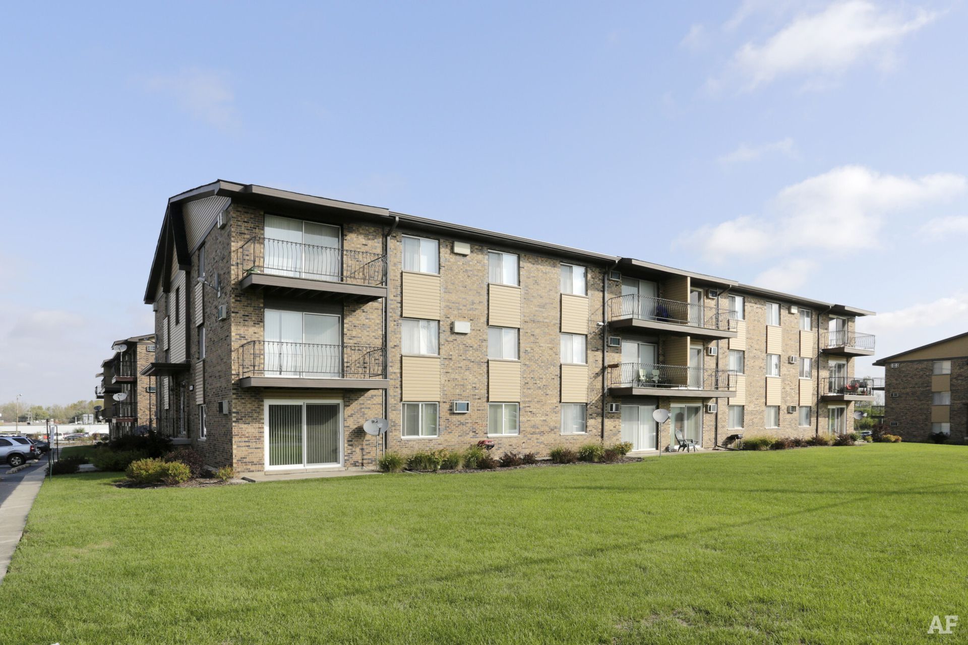 Apartment building with brick and siding exterior, balconies, and a large green lawn on a sunny day.