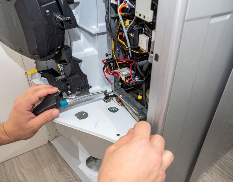 Hands repairing the inside of a silver appliance using a screwdriver, revealing wires and components.